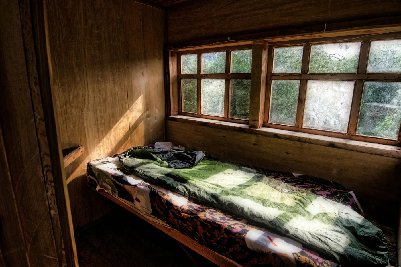 Kleine houten kamer met een bed onder twee ramen die uitkijken op het bos