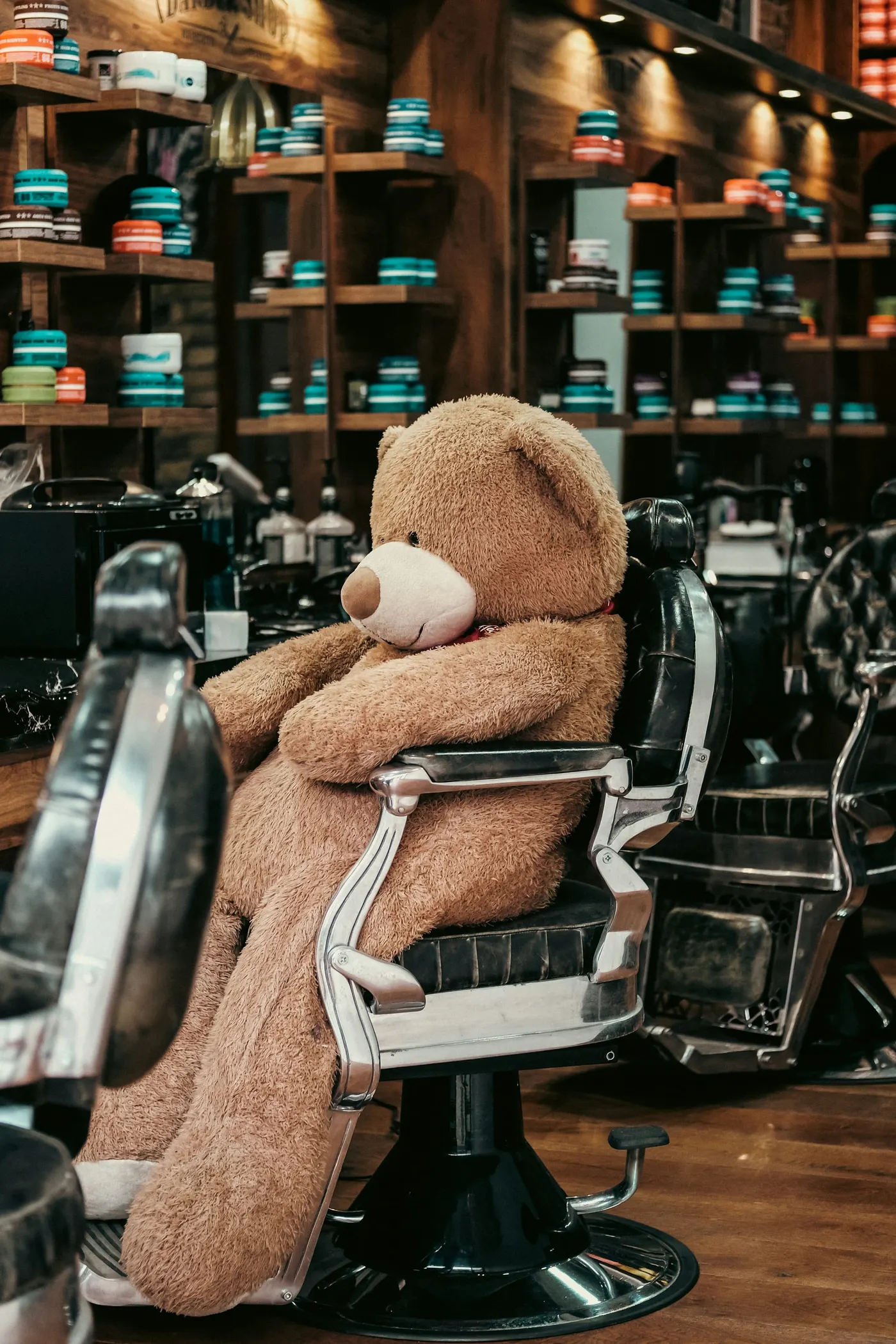Een grote teddybeer zit in een kappersstoel, tussen de schappen met haarproducten